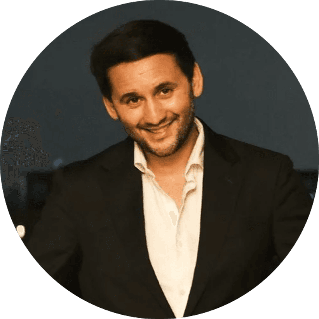 Smiling dark-haired man in a black suit and open-collar white shirt, head-and-shoulders portrait on a dark background.
