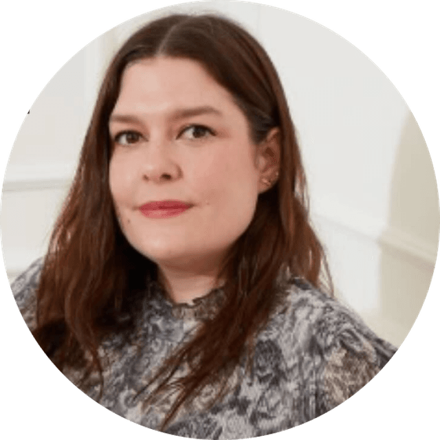 A woman with long brown hair wearing a patterned blouse, standing against a plain white background.