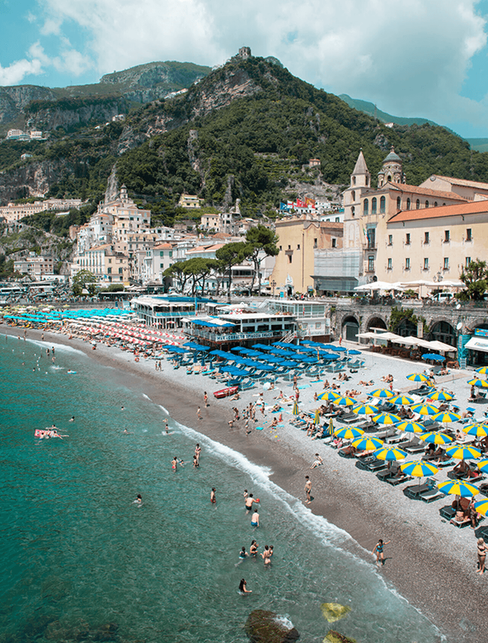 Crowded beach with colorful umbrellas and people swimming in clear blue water, backed by a seaside town and lush green hills.