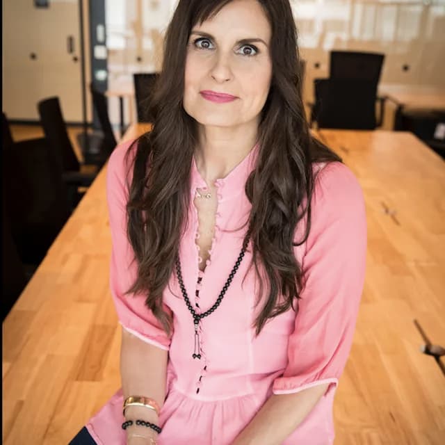 Woman with long brown hair in pink blouse, sitting on a wooden table in a bright conference room.