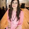 Woman with long brown hair in pink blouse, sitting on a wooden table in a bright conference room.
