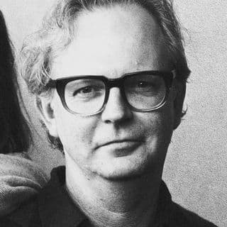 Man with glasses and wavy hair, wearing a dark shirt, looks directly at the camera in a black and white photo.