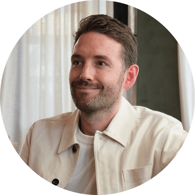 Smiling man with short hair and beard, wearing a light jacket and white shirt, seated indoors with sheer curtains in the background.