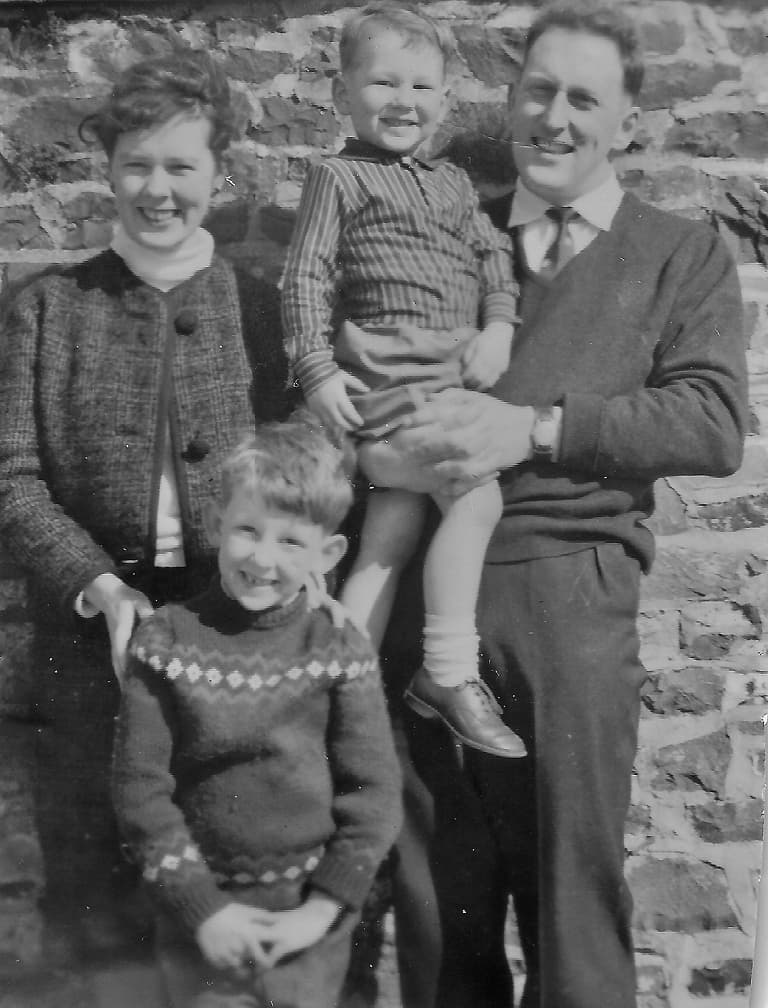 A smiling family of four poses in front of a stone wall. The father holds a young child, and the mother stands beside a boy in a patterned sweater.