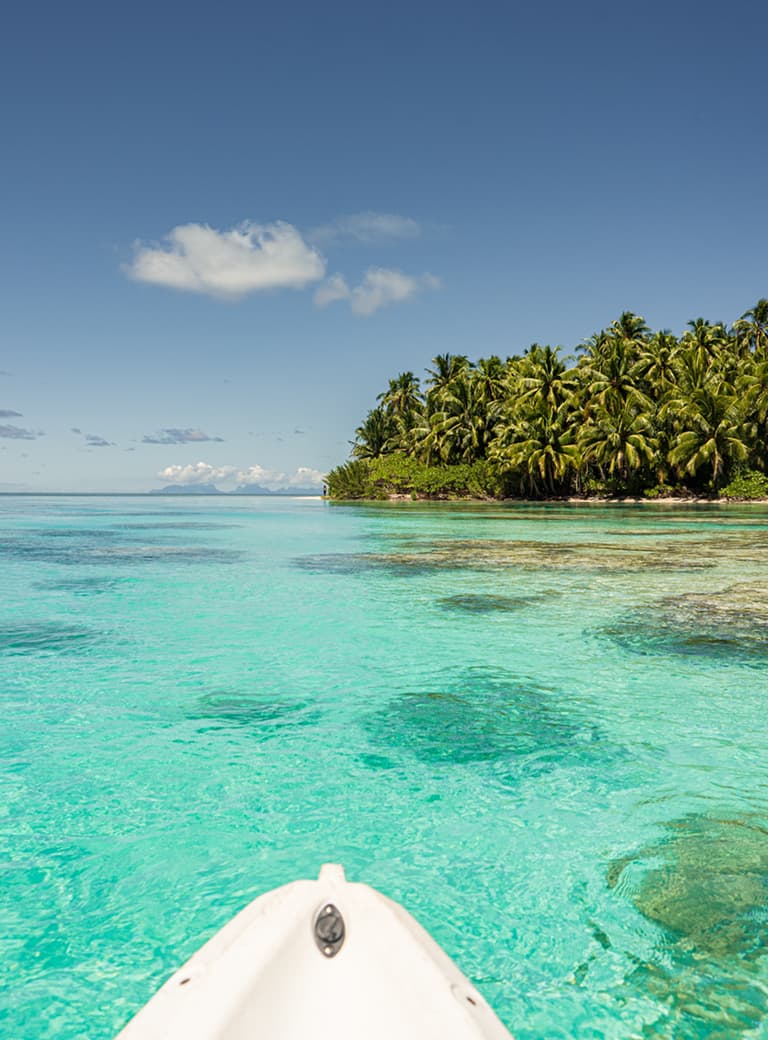 A small boat on clear turquoise water near a tropical island with lush palm trees under a blue sky.