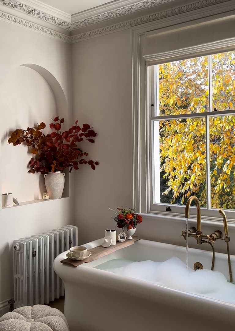 A cozy bathroom with a bubble-filled bathtub, golden faucet, and a large window showing autumn leaves. A vase with red leaves sits on a shelf.