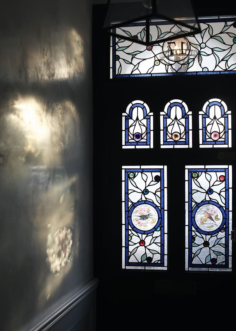 Ornate stained glass door with blue, red, and clear sections. Sunlight casts colorful reflections on the adjacent wall.