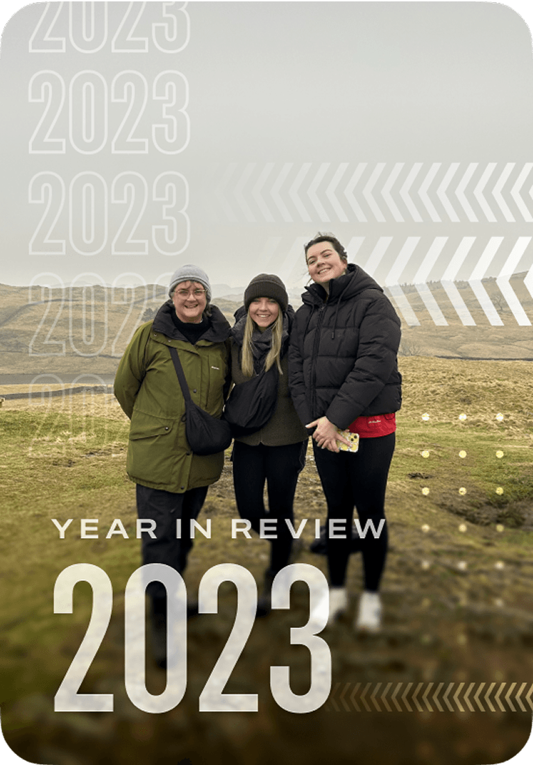 Three people in winter clothing stand smiling on a grassy landscape with hills in the background. Text overlays read "Year in Review 2023."
