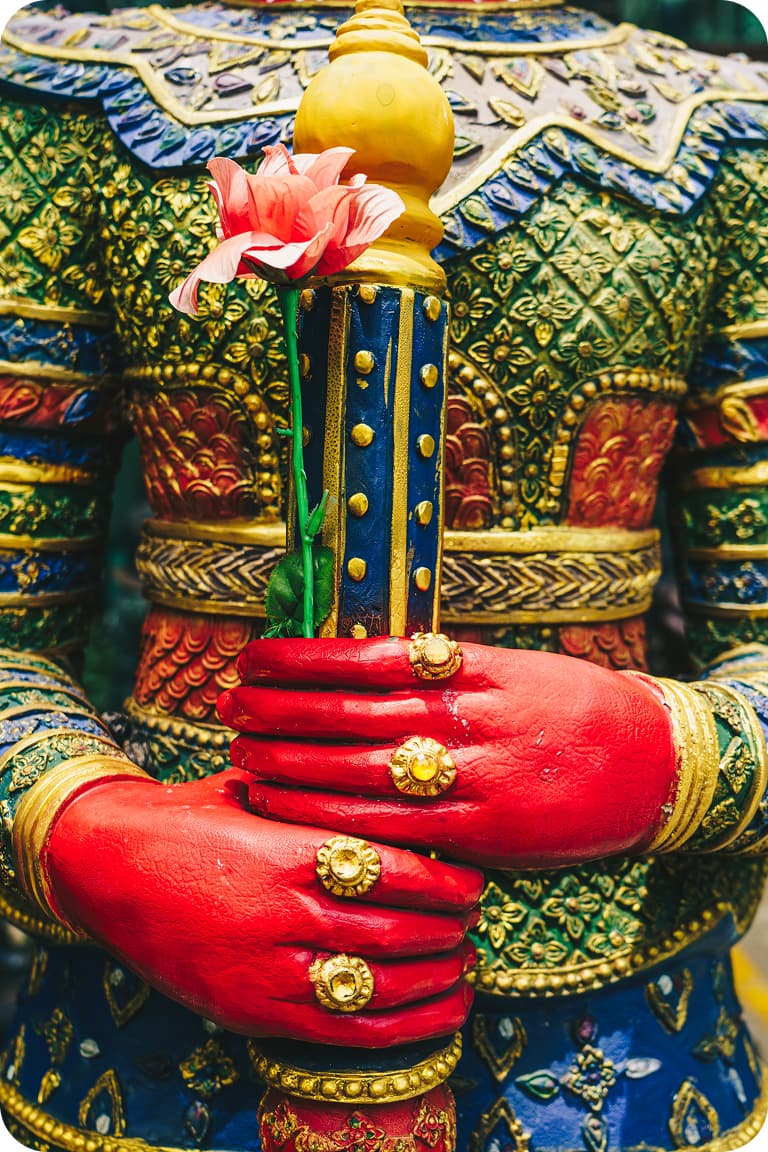 Ornate multicolored statue with red hands wearing gold rings clasping a blue-and-gold staff topped by a pink lotus