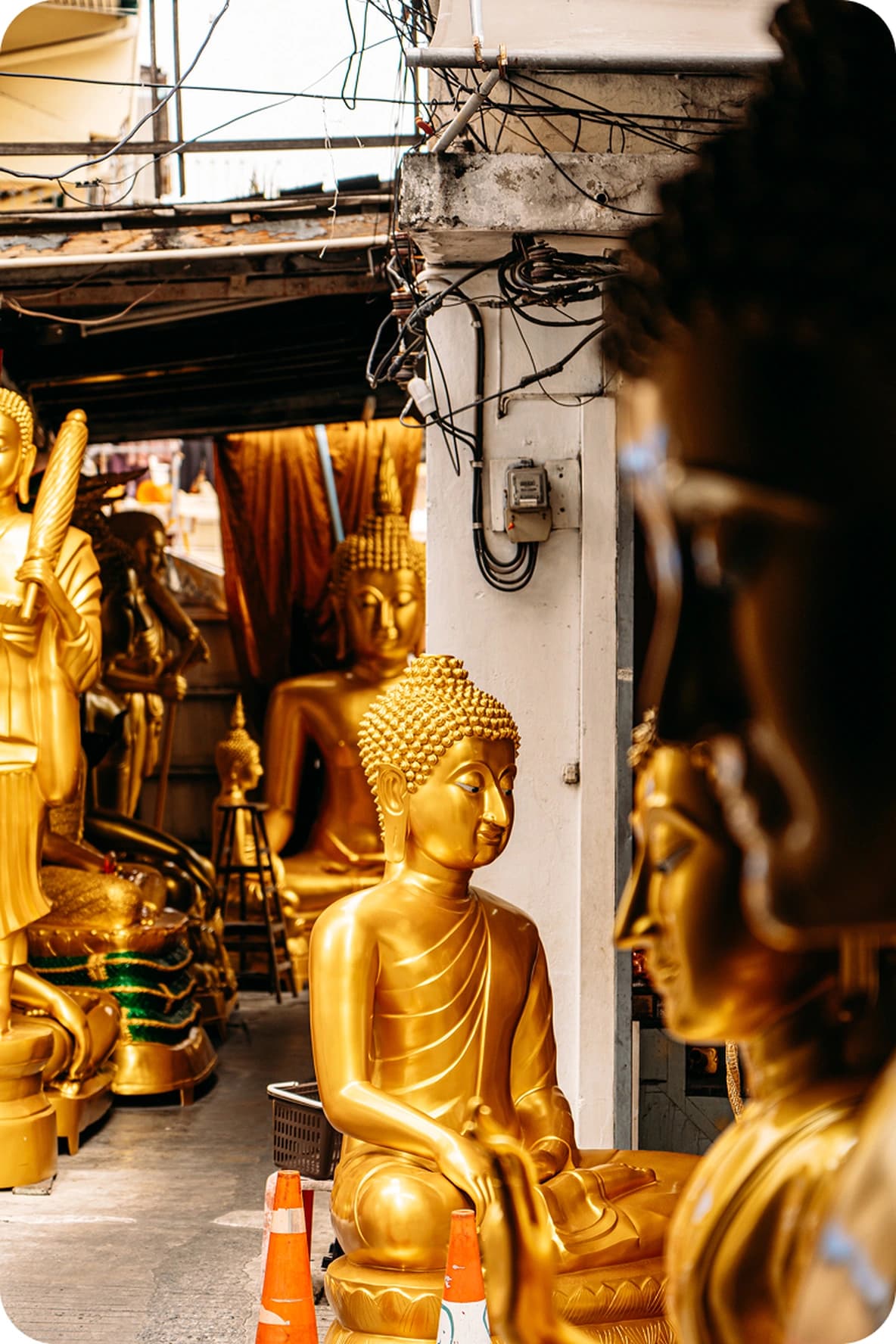 Row of golden Buddha statues in a narrow workshop aisle, several seated figures with traffic cones and visible electrical wiring.