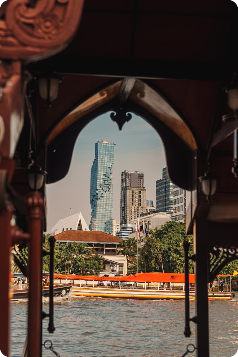 Ornate wooden arch framing river boats with orange canopies and a skyline of modern glass skyscrapers.