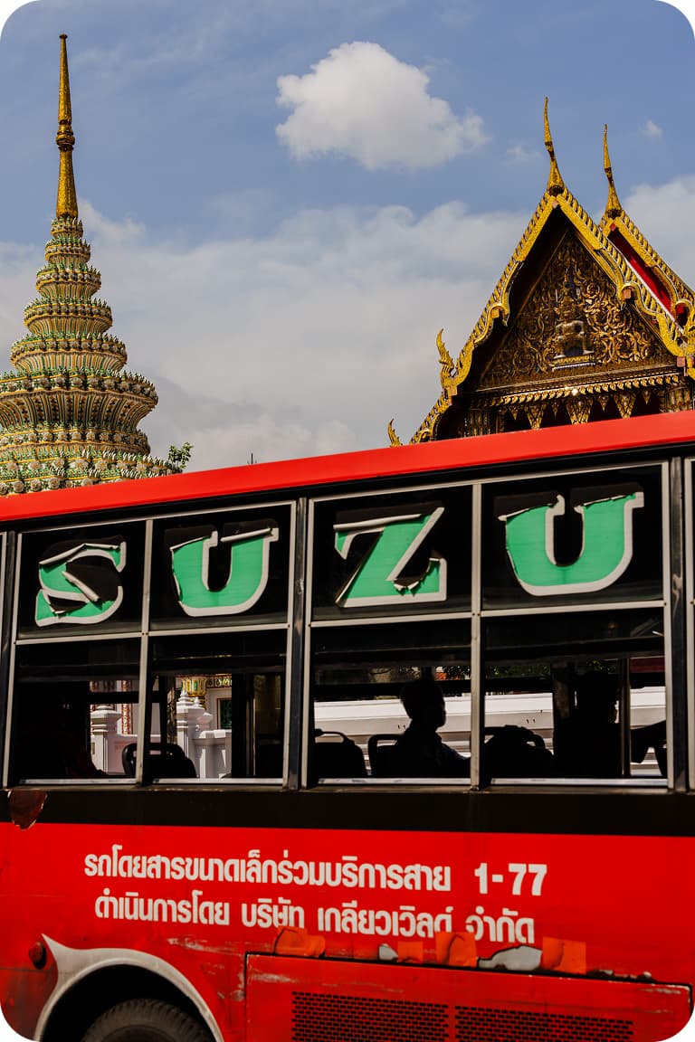 Red city bus with large SUZU letters and passenger silhouettes in front of ornate Thai temple spires under a blue sky.