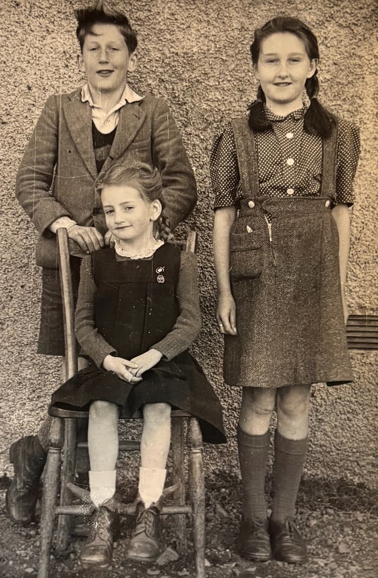 Three children in vintage attire pose against a textured wall; two stand while one sits on a wooden chair, all smiling slightly.