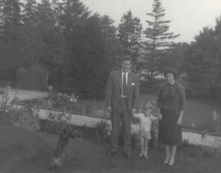 A black and white photo of a family with two adults, two children, and a dog, standing on grass, with trees in the background.