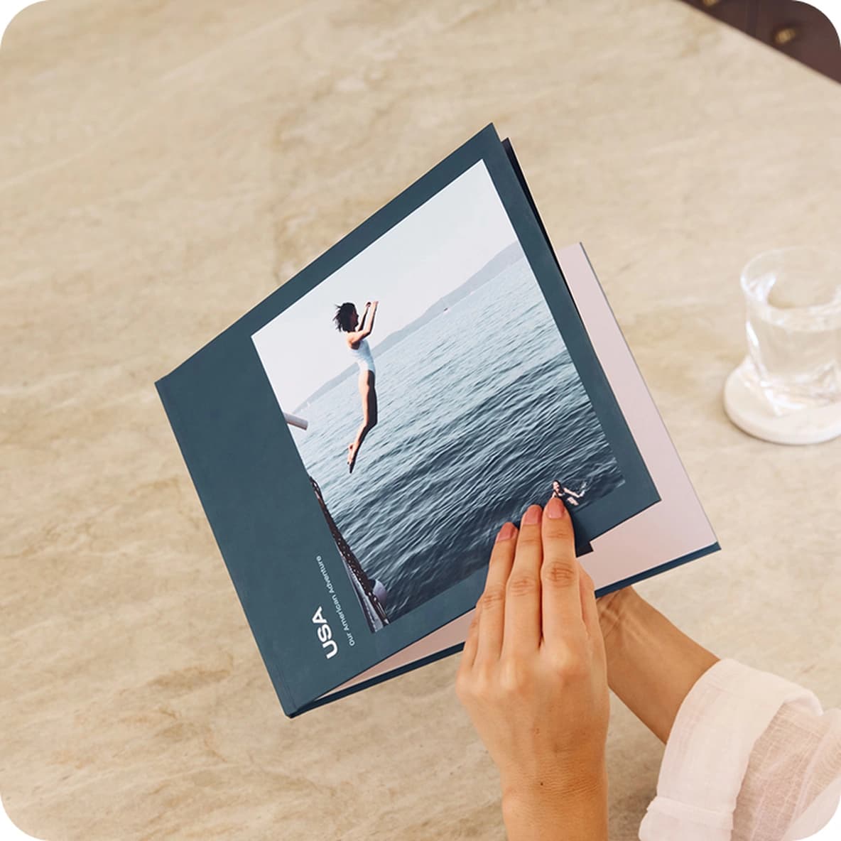Person flipping through a photo book on a marble table, featuring an image of a person diving into the sea. A glass is nearby.
