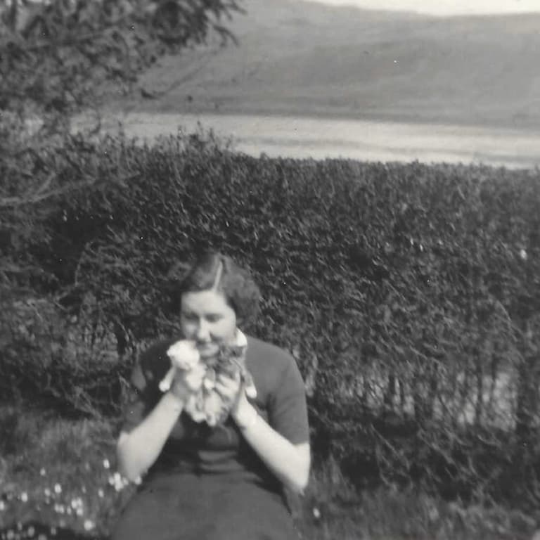 A woman sits outdoors near hedges and a body of water, holding flowers close to her face, with hills in the background. Image is black and white.