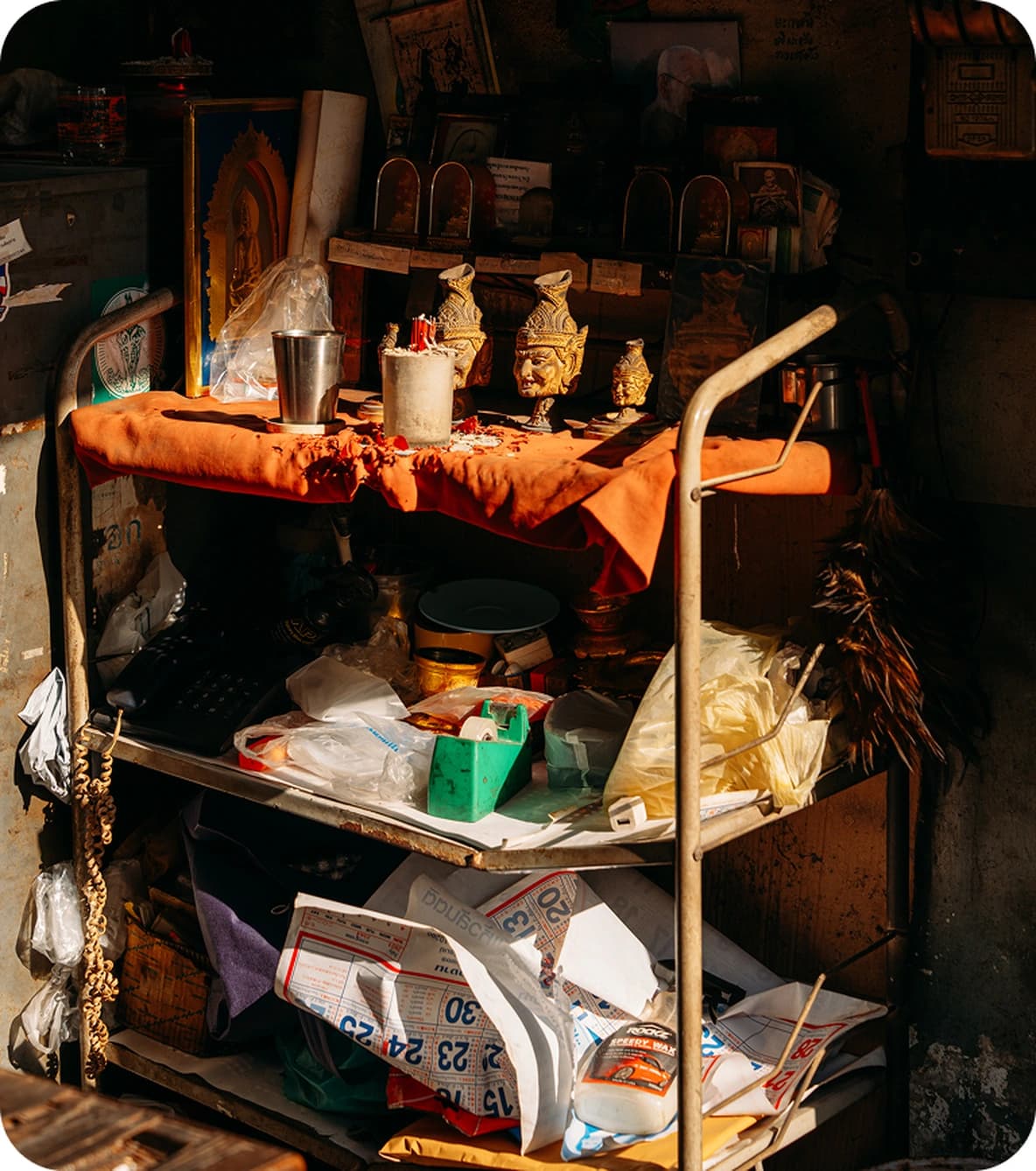 Cluttered metal cart shrine with three brass idols, candles, cups, orange cloth, scattered papers and plastic bags.