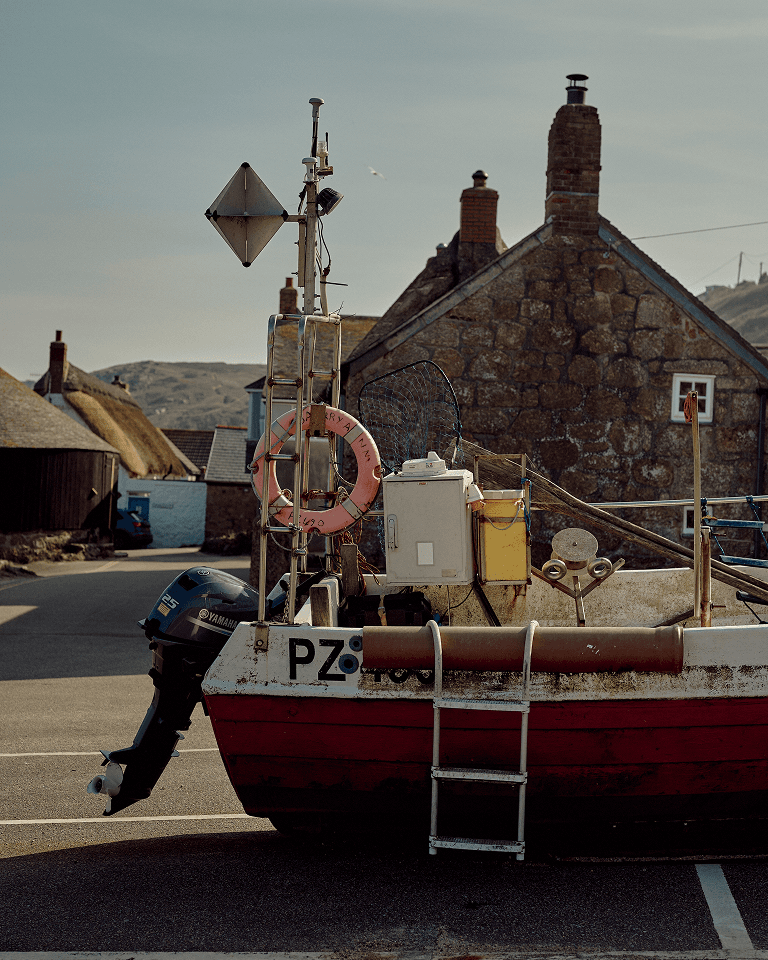 A small red and white fishing boat parked on land with a stone building, chimney, and hills in the background.