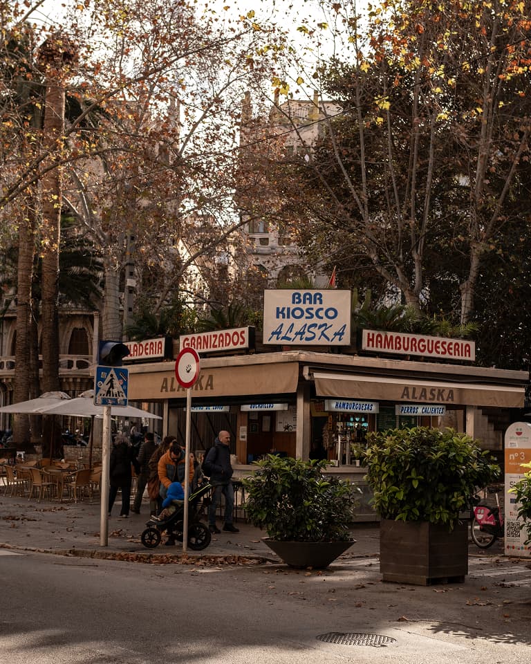 Outdoor kiosk named "Bar Kiosco Alaska" with "Hamburgueseria" and "Granizados" signs, surrounded by trees and people.