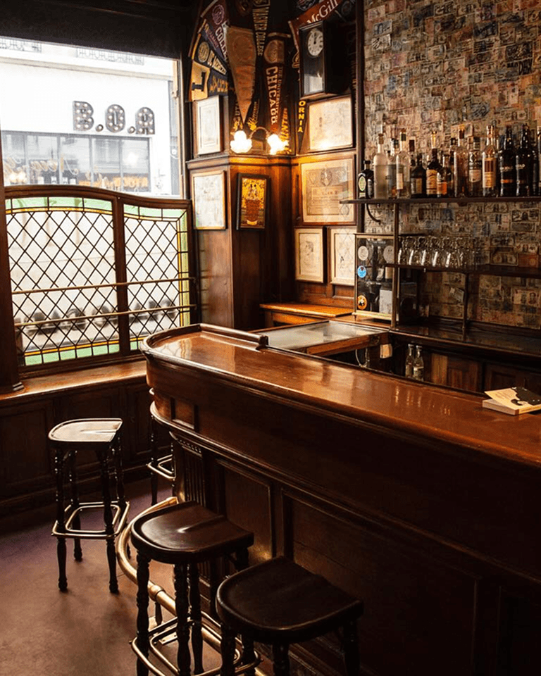 A cozy vintage pub interior with wooden bar, stools, framed memorabilia on walls, and bottles on a shelf, lit by warm overhead lights.