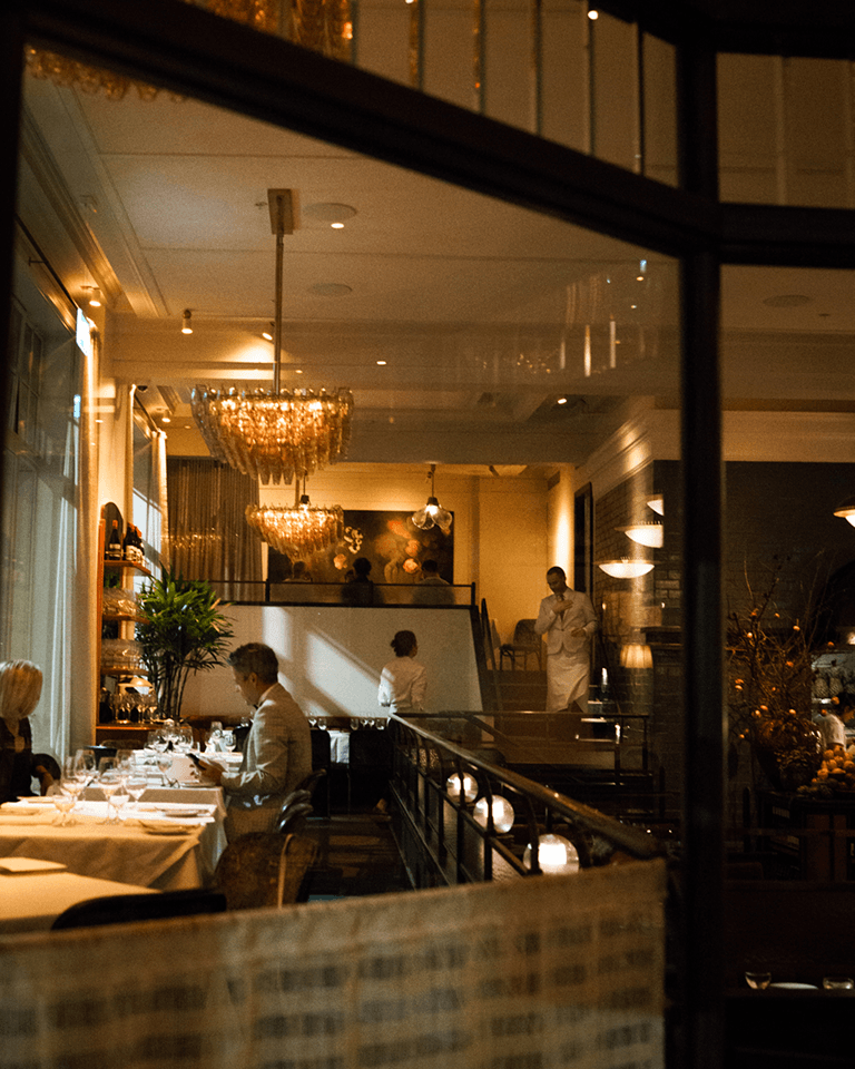 Elegant restaurant interior with dim lighting, chandeliers, and patrons dining at candlelit tables. A waiter stands near the stairs.