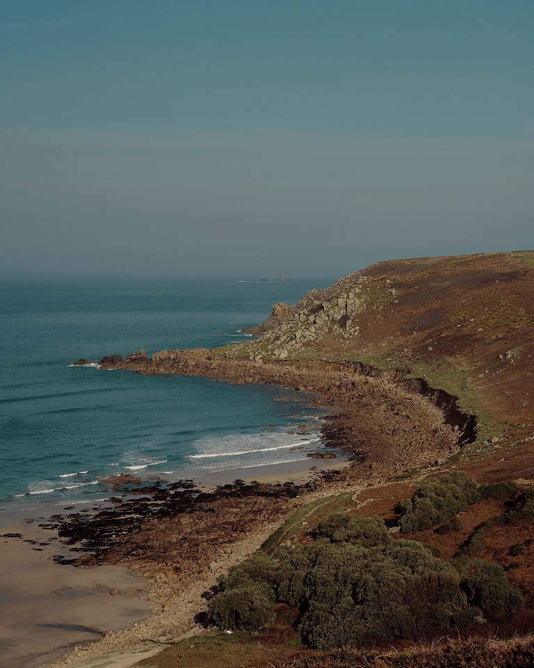 A rugged coastline with rocky cliffs and a sandy beach bordered by green vegetation under a clear blue sky.