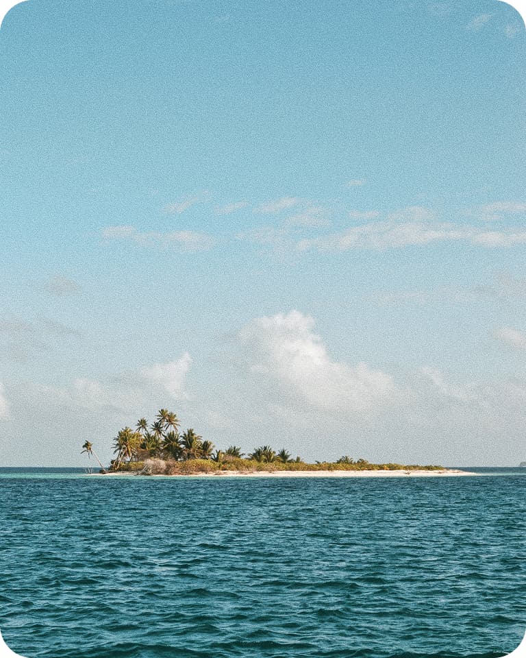 Tiny tropical island with palm trees and sandy shore surrounded by calm blue ocean under clear sky.