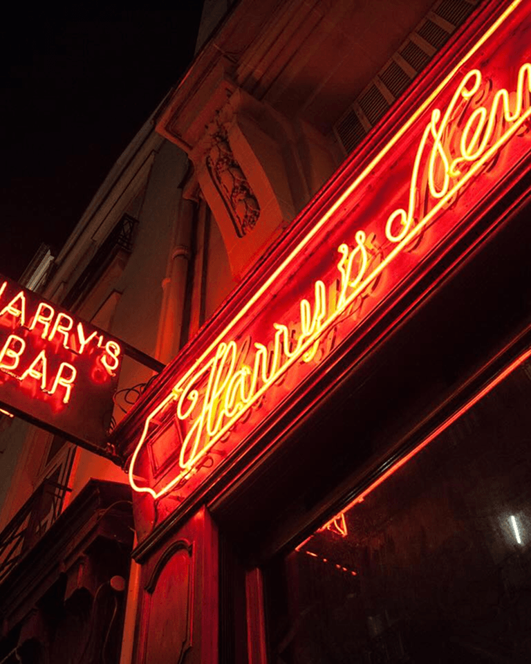 Bright red neon signs reading "Harry's Bar" glow against a dark night sky, mounted on a classic building facade.