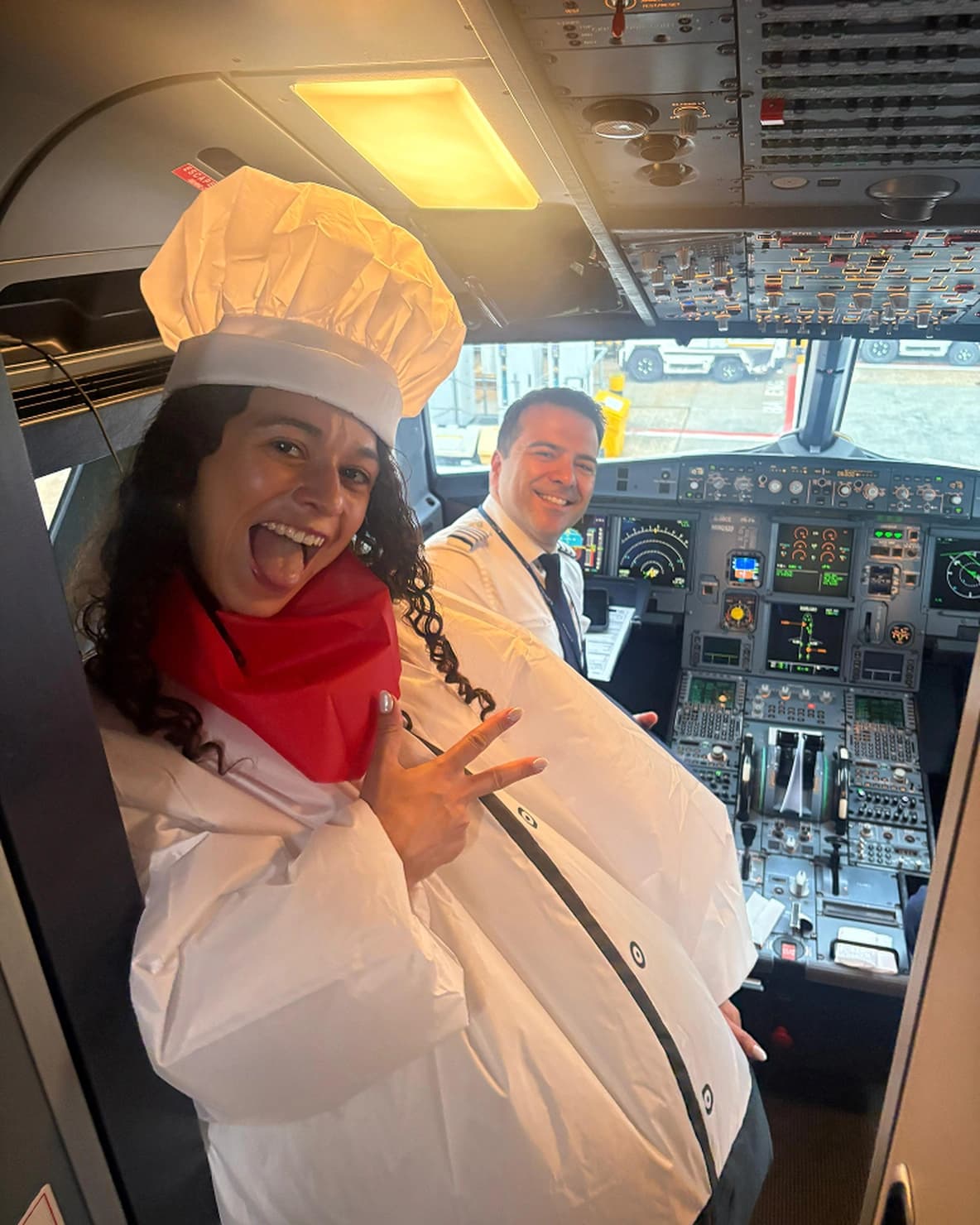 Woman in an inflatable chef costume making a peace sign in an airplane cockpit, with a smiling pilot in uniform behind her.