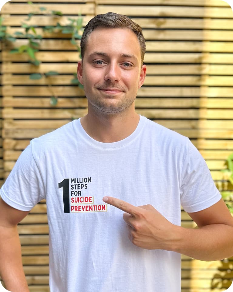 Person wearing a white T-shirt with "1 Million Steps for Suicide Prevention" text, pointing at the message against a wooden slat background.