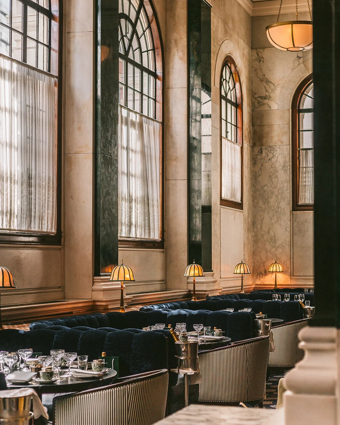 Elegant dining area with tall arched windows, blue velvet seating, marble walls, and vintage table lamps creating a cozy ambiance.