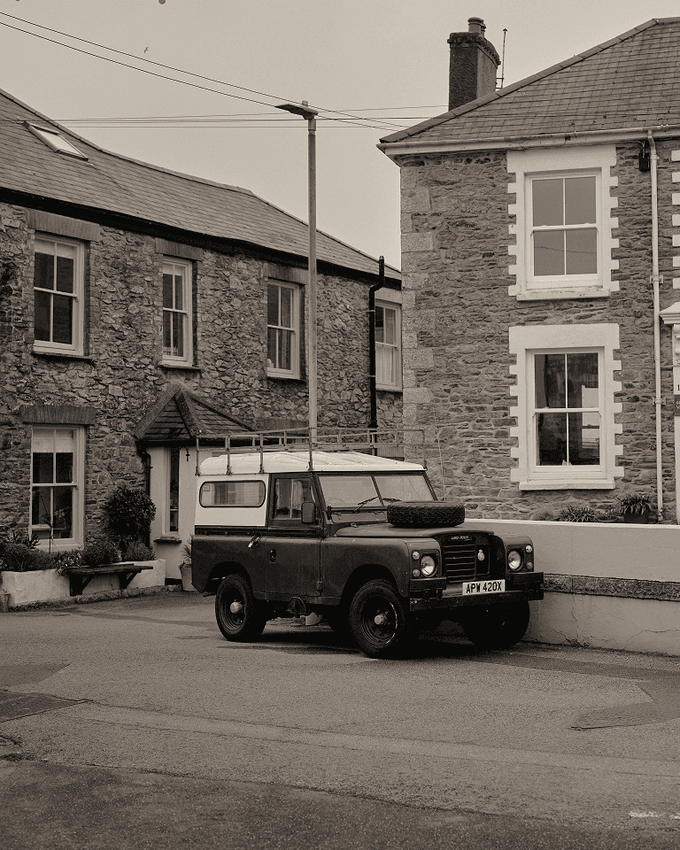 A vintage Land Rover parked on a quiet street in front of two stone houses with white-trimmed windows.