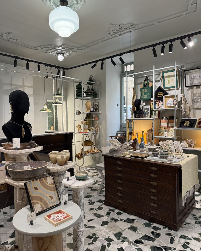 A boutique store displays various home decor items and accessories on shelves and tables, with patterned flooring and elegant lighting.