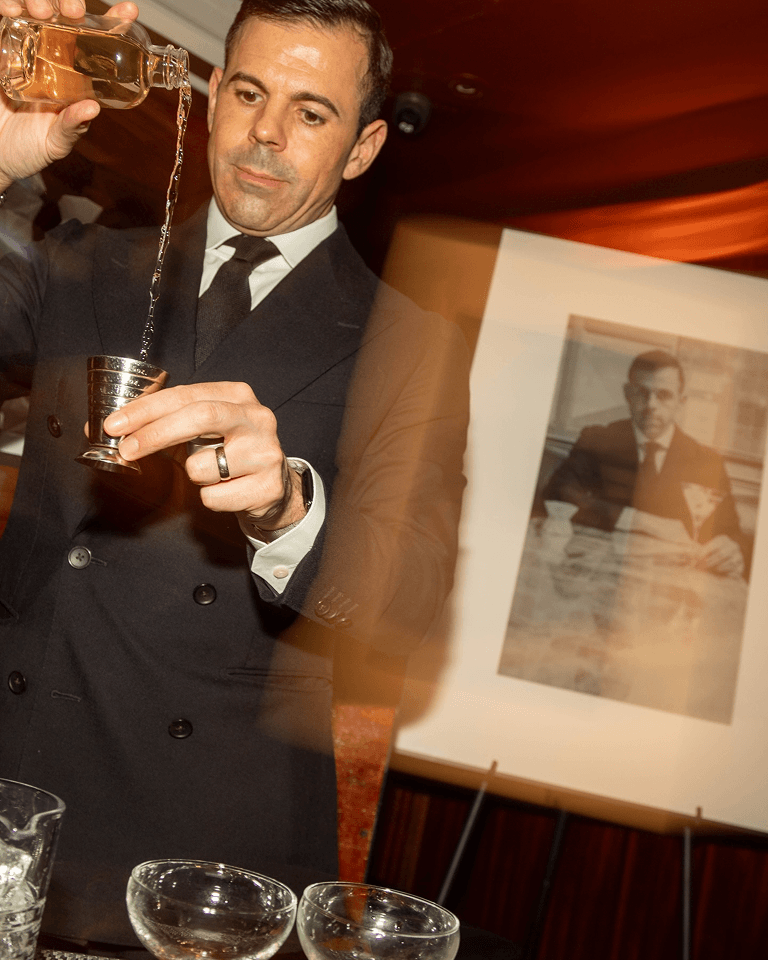 A man in a suit pours a drink into a jigger, with a blurred vintage photo in the background.