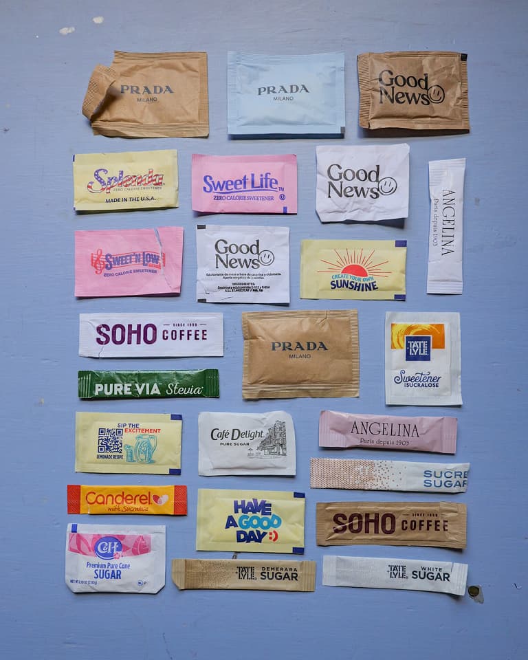 Grid of 25 assorted sugar and sweetener packets in various colors and brands, neatly arranged on a pale blue wooden surface.