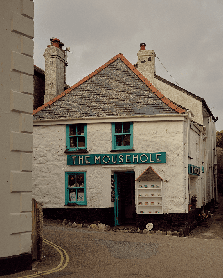 Old stone building with "The Mousehole" sign, teal window frames, and overcast sky; small shop located on a narrow street corner.