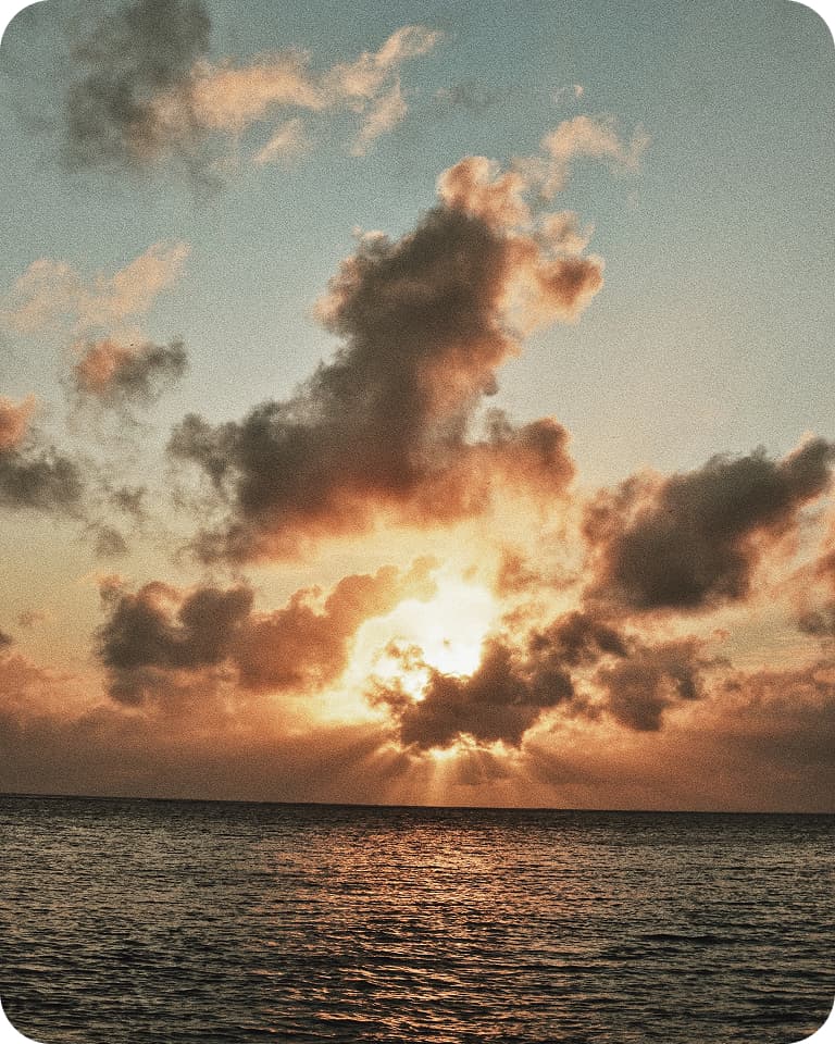 Sunset over the ocean with golden sun rays breaking through dramatic clouds and reflecting on rippled water.