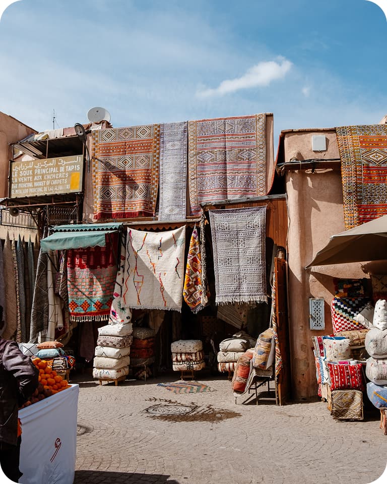 Street market with colorful rugs and textiles hanging on a building, surrounded by pillows and baskets under a clear blue sky.
