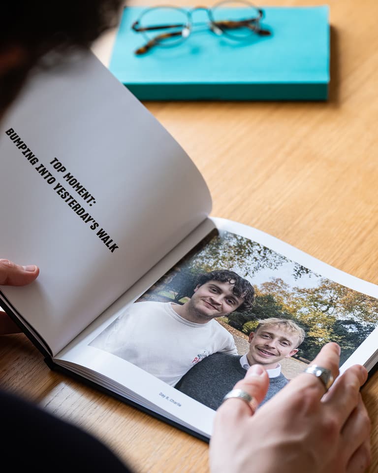 Person holding an open book with a photo of two smiling individuals and text: "Top Moment: Bumping Into Yesterday's Walk." Blue item and glasses in the background.