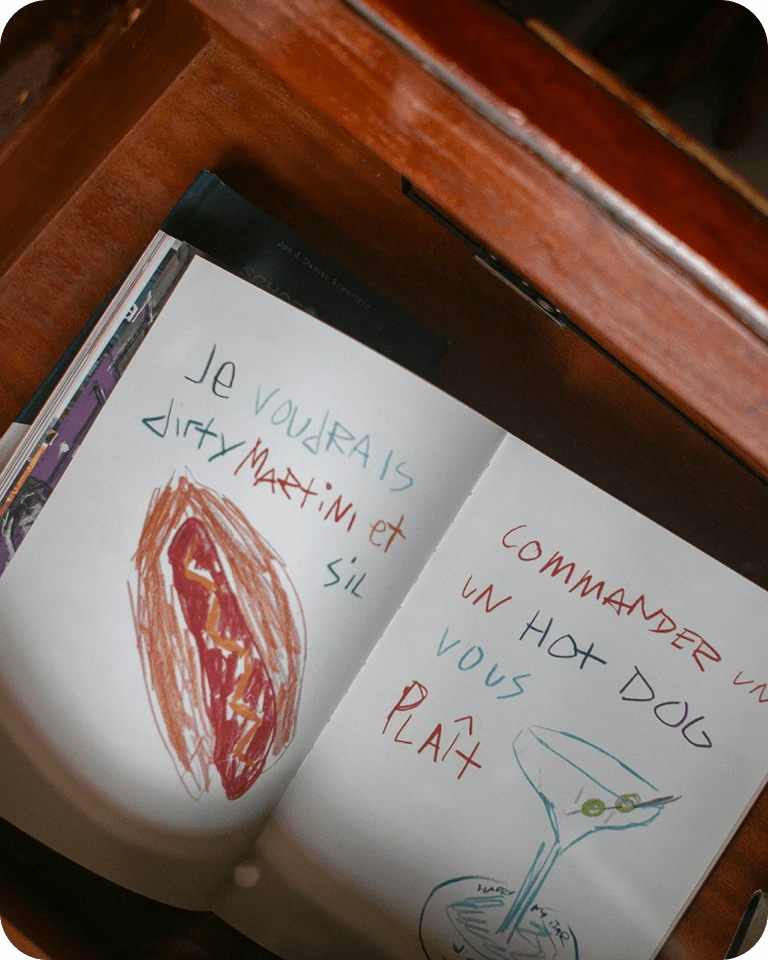 Open notebook in a drawer with colorful text in French and drawings of a hot dog and a martini glass.