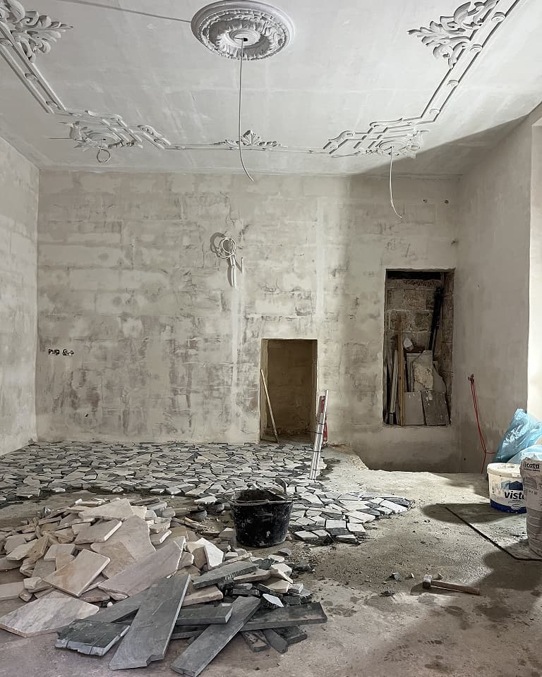 Room under renovation with unfinished walls, ornate ceiling, scattered tiles, and construction materials. A ladder leans against the wall.