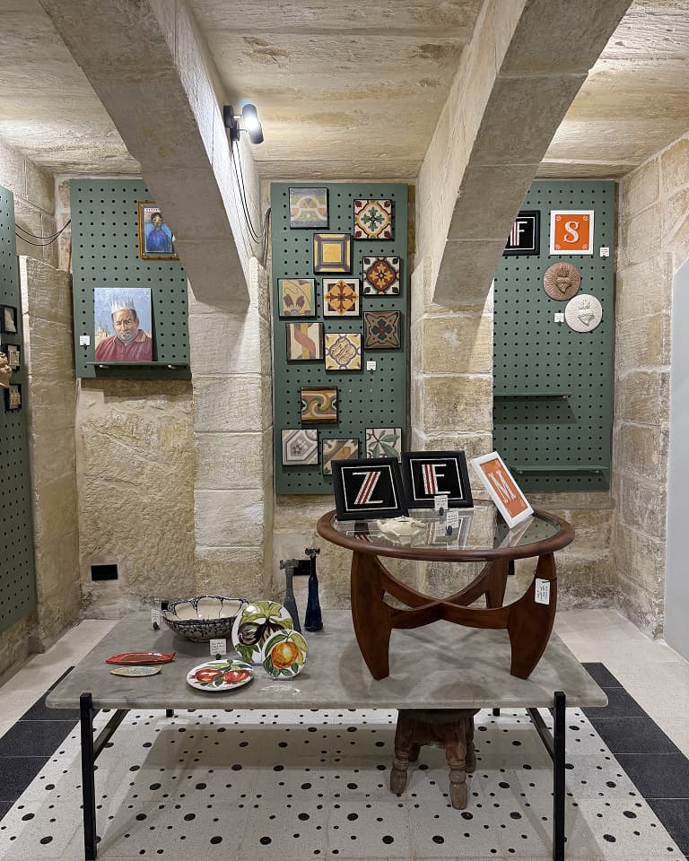 Art gallery room with stone walls, displaying framed paintings, tiled artwork on pegboards, and a central wooden table with books and decorations.