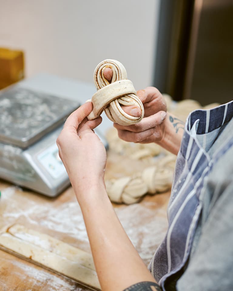 Person shaping a piece of dough in a kitchen, with a weighing scale and more dough on the floured surface in the background.