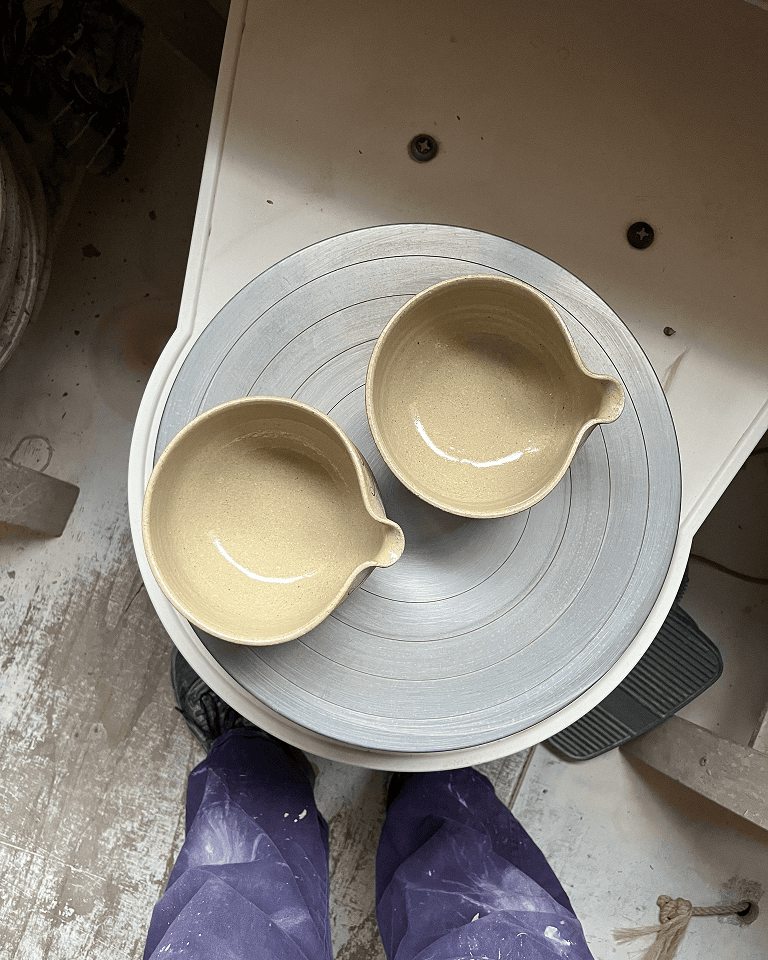 Two beige ceramic bowls with spouts sit on a metal pottery wheel. Below, purple pants and black shoes are visible on a dusty floor.