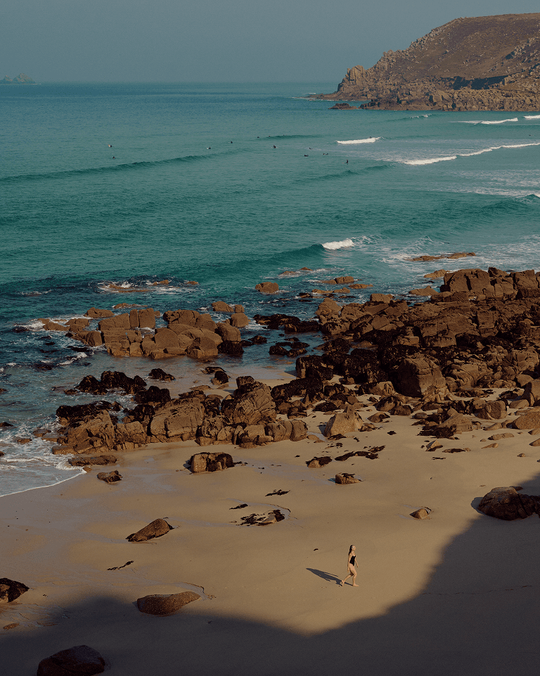 A person walks on a rocky beach with turquoise waves, under a clear sky, beside cliffs in the background.