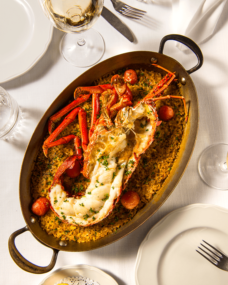 A lobster dish with rice and vegetables, served in a brass oval pan on a white tablecloth, surrounded by wine glasses and empty plates.