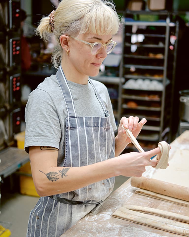 Person with glasses and striped apron shaping clay in a pottery studio, surrounded by tools and pottery materials.