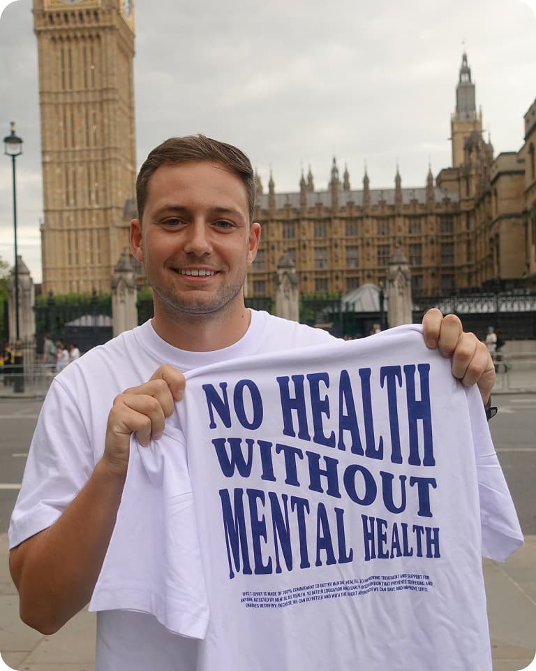 Man holding a shirt with "No Health Without Mental Health" text, standing in front of the Houses of Parliament.