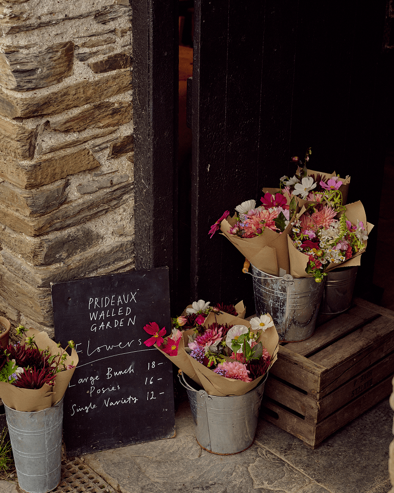 Buckets of colorful flowers in brown paper wraps sit outside a dark wooden door, with a sign for "Prideaux Walled Garden" listing flower prices.