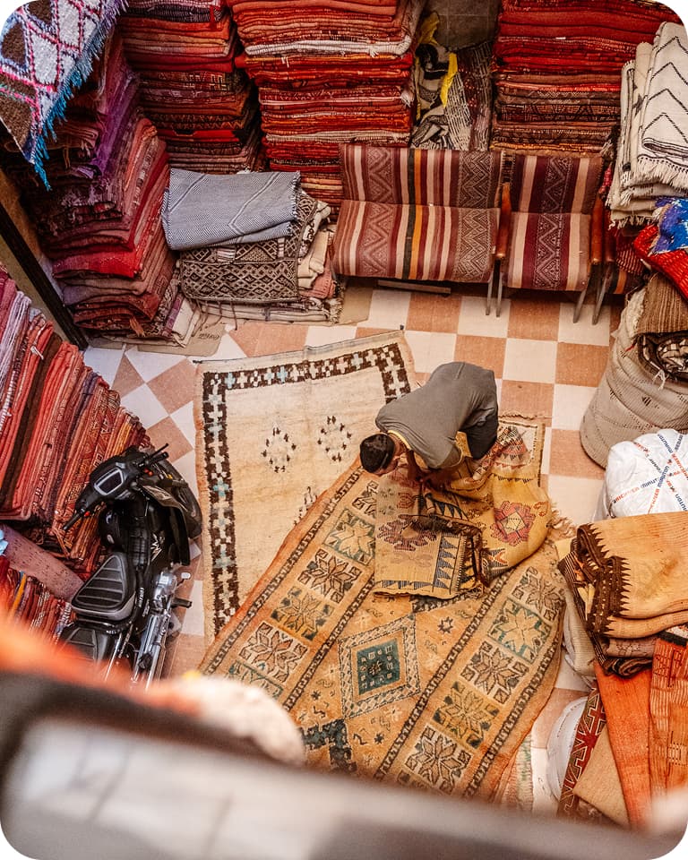 A person examines a large, patterned rug in a shop filled with stacked colorful textiles and carpets, viewed from above.